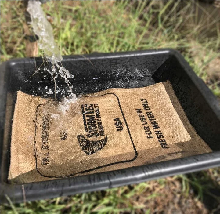 Sac en jute STORMBAG posé dans une benne noire, l’eau tombe dessus; étiquette imprimée mentionnant « Stormbag », usage, et date; produit carton de 10 sacs.
