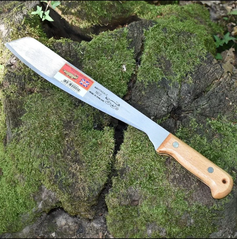 Couteau long à lame argentée et manche en bois clair, posé sur de la mousse verdoyante et un tronc; étiquette sur la lame.