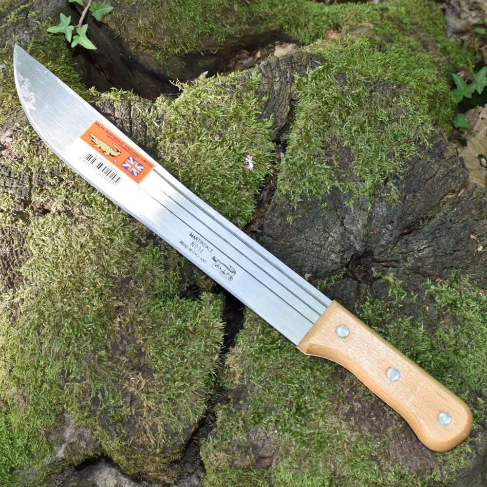Couteau de survie pliant à lame épaisse et longue, avec manche en bois clair; posé sur faible mousse et bois, étiquette orange sur la lame.