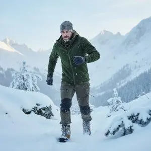 Homme en tenue de outdoor en hiver, veste verte, pantalon et bottes sur terrain enneigé avec montagnes en arrière-plan.