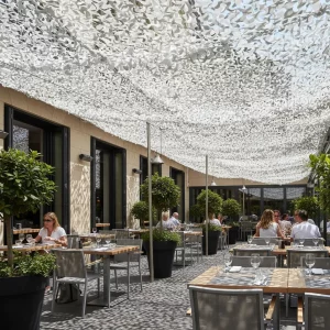 Terrasse extérieure ombragée par un filet d’ombrage blanc 5x5m, tables en bois, chaises grises et plantes en pots, ambiance restaurant en journée.