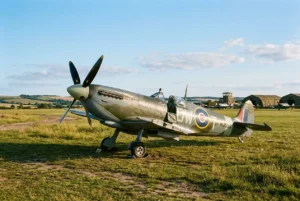 Avion de chasseVintage sur herbe, couvercle d’ailes et train d’atterrissage, décor champêtre et hangars au loin, représentant les avions militaires de légende.