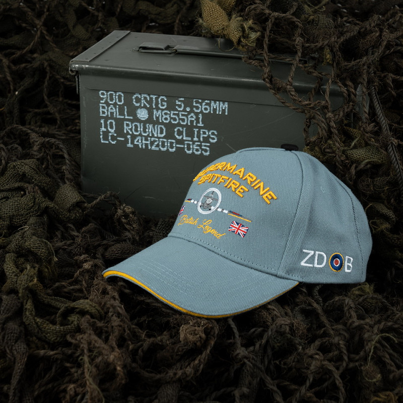 Casquette RAF Supermarine Spitfire bleu avec inscriptions jaunes et drapeau britannique, posée près d’une caisse militaire.